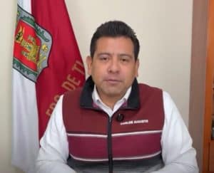 Persona en una oficina con bandera de Tlaxcala al fondo, usando un chaleco con su nombre, en un entorno formal y profesional.