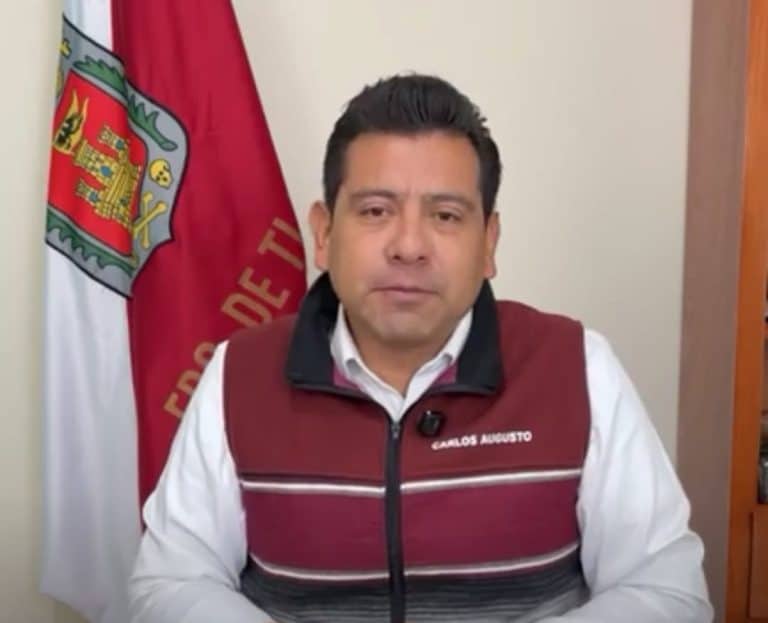 Persona en una oficina con bandera de Tlaxcala al fondo, usando un chaleco con su nombre, en un entorno formal y profesional.