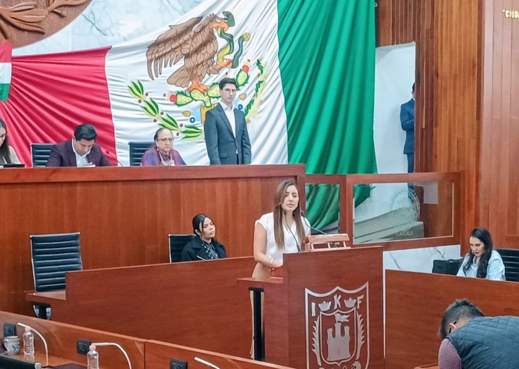 Persona hablando en un acto oficial en una sala con bandera de México de fondo y otros asistentes presentes.