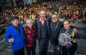 Un grupo de cinco músicos en un concierto en vivo, con una multitud de espectadores en el fondo, en un evento cultural en Tlaxcala, México.