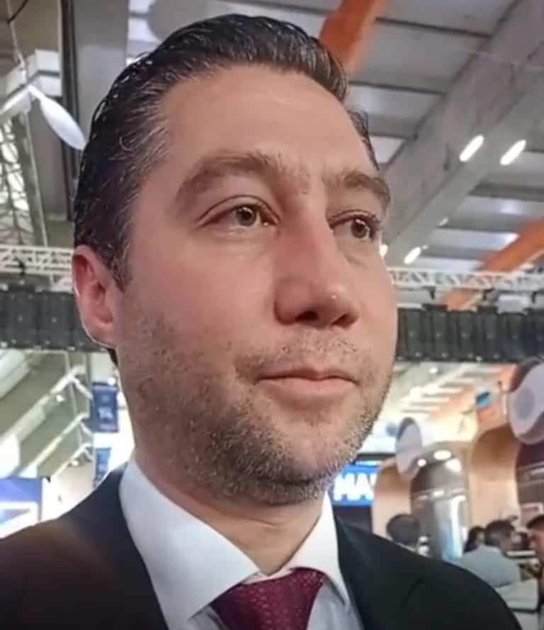Hombre con cabello oscuro peinado hacia atrás, usando traje negro, camisa blanca y corbata burdeos, en un entorno moderno y bien iluminado, posiblemente en un aeropuerto o centro comercial.