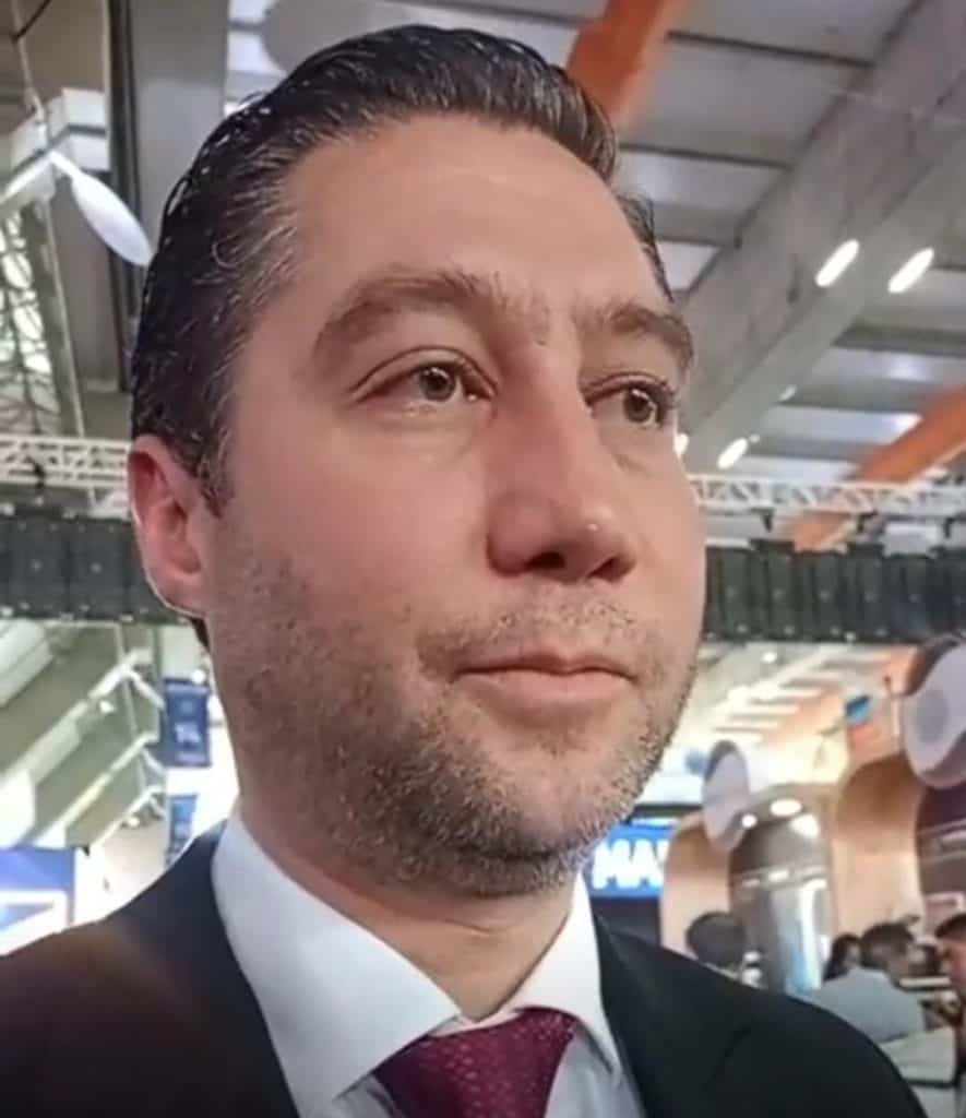Hombre con cabello oscuro peinado hacia atrás, usando traje negro, camisa blanca y corbata burdeos, en un entorno moderno y bien iluminado, posiblemente en un aeropuerto o centro comercial.