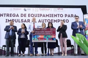 Grupo de personas en un evento de entrega de equipo para impulsar el autoempleo en Tlaxcala, con un cartel que destaca la importancia de maquinaria y herramientas para negocios locales.