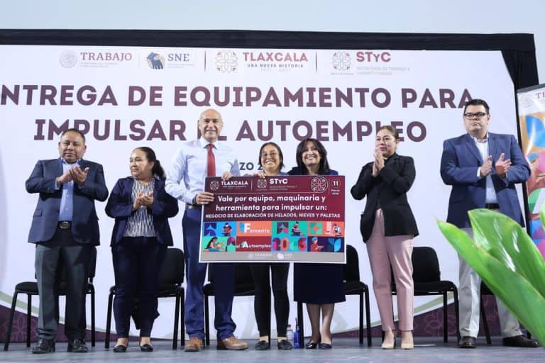 Grupo de personas en un evento de entrega de equipo para impulsar el autoempleo en Tlaxcala, con un cartel que destaca la importancia de maquinaria y herramientas para negocios locales.
