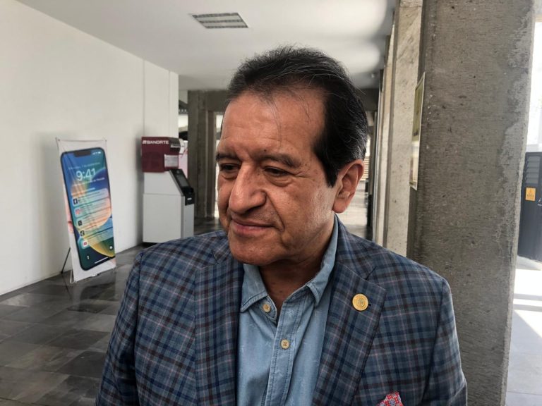 Hombre mayor con traje a cuadros y camisa azul, en un espacio interior con paredes blancas y una estructura de concreto, en un entorno que parece ser un edificio público o una oficina.