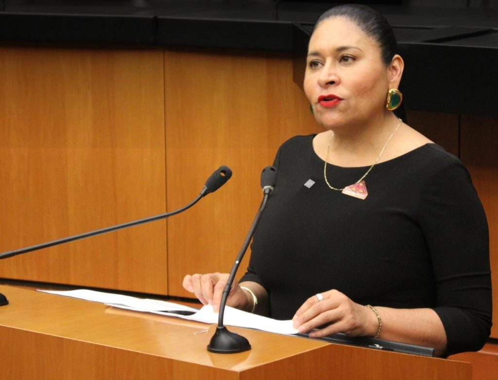 Mujer en conferencia, hablando frente a micrófono en un entorno formal, con fondo de madera y vestimenta profesional negra.