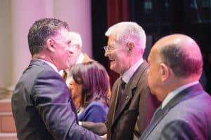 Hombre y mujer mayores saludándose y sonriendo en un evento formal, con otras personas en el fondo.
