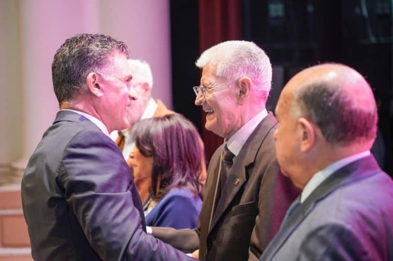 Hombre y mujer mayores saludándose y sonriendo en un evento formal, con otras personas en el fondo.
