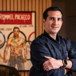 Persona con uniforme de entrenador de tiro con arco en un evento deportivo en Tlaxcala, con fondo de cartel de los Juegos Olímpicos y una atleta en el fondo.