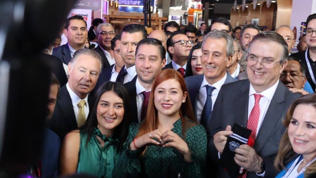 Junta de políticos y líderes en un evento público, con sonrisas y gestos de camaradería, en un ambiente formal y festivo, con varias personas en el fondo y luces brillantes.