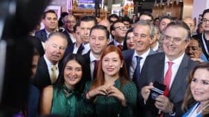 Junta de políticos y líderes en un evento público, con sonrisas y gestos de camaradería, en un ambiente formal y festivo, con varias personas en el fondo y luces brillantes.
