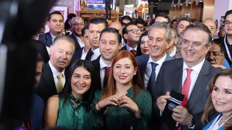 Junta de políticos y líderes en un evento público, con sonrisas y gestos de camaradería, en un ambiente formal y festivo, con varias personas en el fondo y luces brillantes.