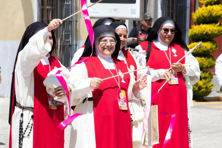 Grupo de religiosas vestidas con hábito y capa blanca y roja, celebrando con alegría y banderas en mano en una calle de Tlaxcala durante una festividad religiosa o evento comunitario.