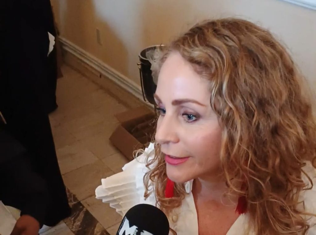Asistente de noticias de Tlaxcala en entrevista, mujer con cabello rizado y expresión seria, sosteniendo micrófono en interior de oficina o sala de reuniones, fondo con pared y objetos decorativos.