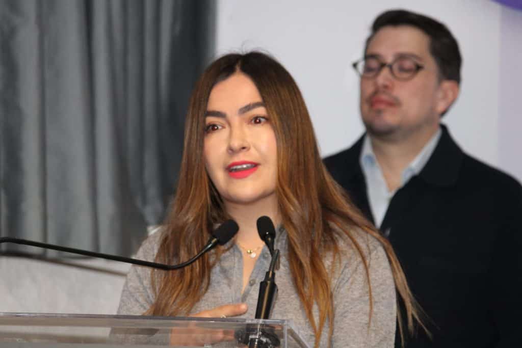 Mujer hablando en un evento formal con micrófono y fondo de cortinas grises, en un entorno profesional.