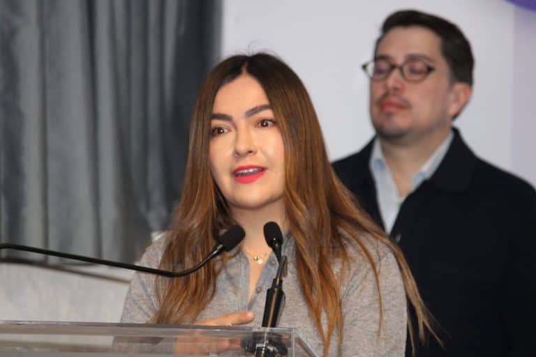 Mujer hablando en un evento formal con micrófono y fondo de cortinas grises, en un entorno profesional.