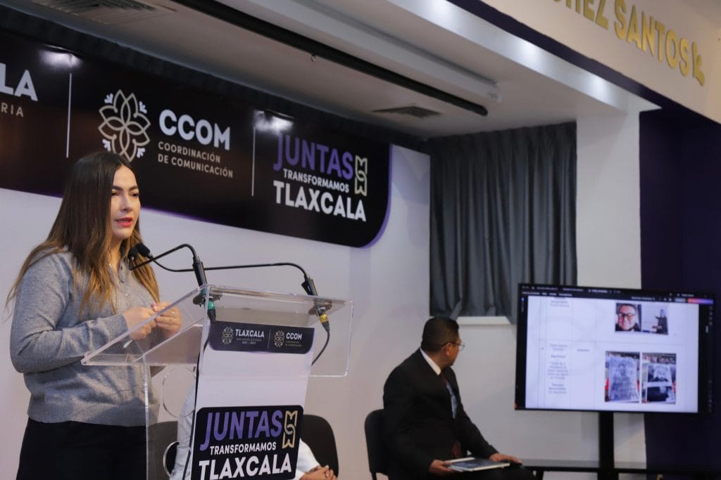 Capta de una mujer hablando en un podio durante una conferencia en un evento oficial en Tlaxcala, con un hombre sentado en una mesa y una pantalla digital mostrando información relevante.