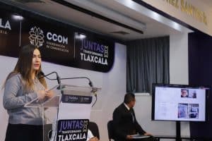 Capta de una mujer hablando en un podio durante una conferencia en un evento oficial en Tlaxcala, con un hombre sentado en una mesa y una pantalla digital mostrando información relevante.