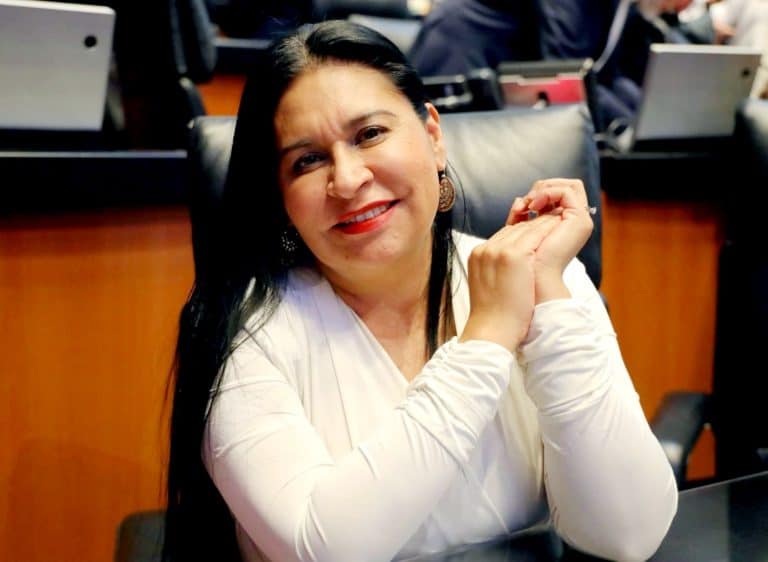 Una mujer sonriendo en un entorno formal, posiblemente en una reunión o conferencia, con fondo de mesas y computadoras.