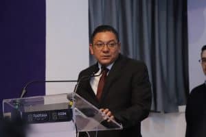 El hombre en traje negro y corbata, con gafas, hablando en un podio durante una conferencia o evento oficial en Tlaxcala, México.
