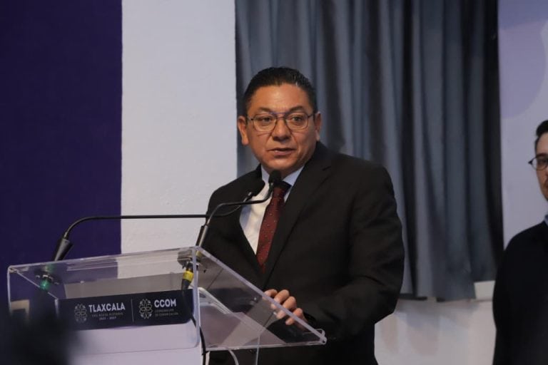 El hombre en traje negro y corbata, con gafas, hablando en un podio durante una conferencia o evento oficial en Tlaxcala, México.