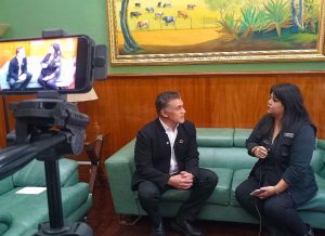 Entrevista en un ambiente formal con dos personas sentadas en un sillón verde, una de ellas con micrófono en mano, en un espacio decorado con un cuadro de animales y plantas en la pared.