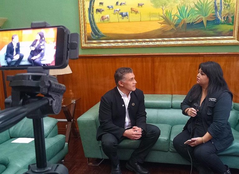 Entrevista en un ambiente formal con dos personas sentadas en un sillón verde, una de ellas con micrófono en mano, en un espacio decorado con un cuadro de animales y plantas en la pared.