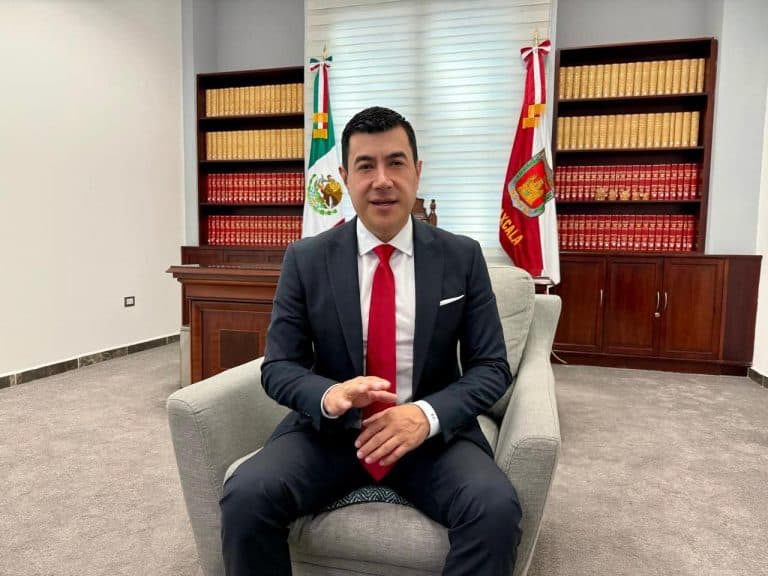 Hombre en traje formal sentado en una sala de oficina con banderas y estanterías llenas de libros en el fondo, en un entorno profesional y oficial.