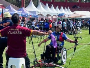 Dispositivo de arco y flecha adaptado para personas con discapacidad en un evento al aire libre, con varias carpas blancas y asistentes en el fondo.
