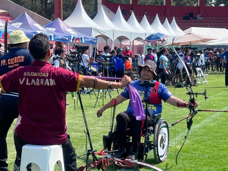 Dispositivo de arco y flecha adaptado para personas con discapacidad en un evento al aire libre, con varias carpas blancas y asistentes en el fondo.