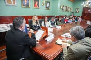 Reunión formal en una sala con varios asistentes, algunos tomando notas y otros escuchando, en un ambiente profesional y decorado con retratos en las paredes.