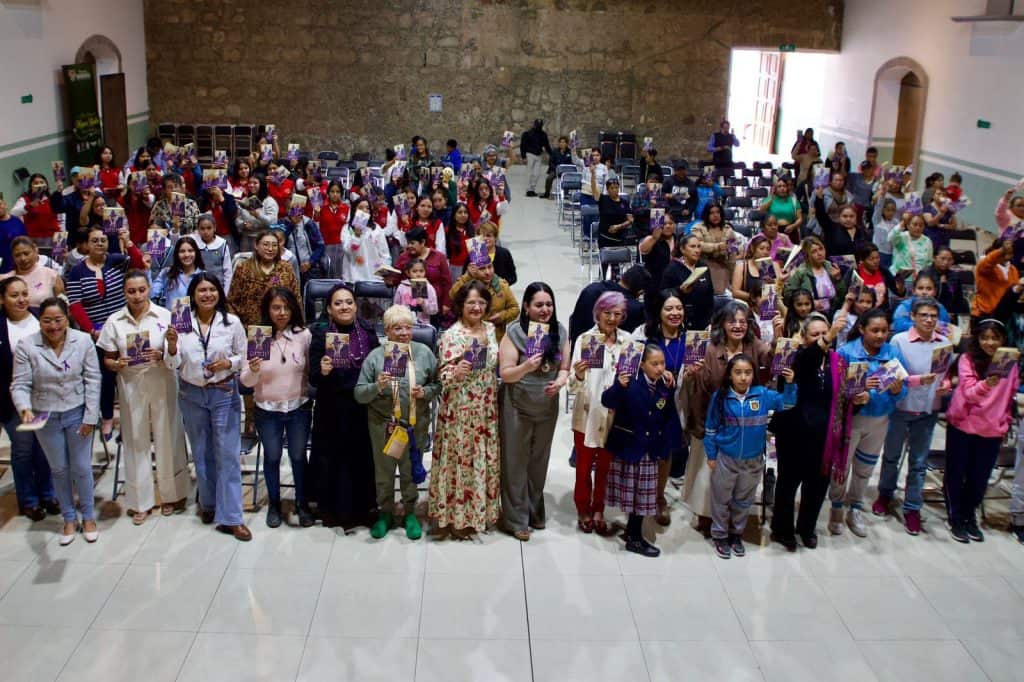 Gran grupo de personas, incluyendo niños y adultos, reunidas en un evento en un salón amplio con paredes de color claro y ventanas grandes que dejan entrar luz natural.