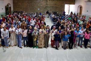 Gran grupo de personas, incluyendo niños y adultos, reunidas en un evento en un salón amplio con paredes de color claro y ventanas grandes que dejan entrar luz natural.