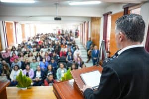 Amplificador de voz en un auditorio lleno de asistentes, vista desde el escenario, con un orador en primer plano usando un dispositivo digital para dirigirse a la audiencia.