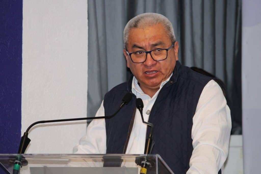 Hombre mayor con gafas y cabello gris, hablando en un micrófono durante una conferencia o evento formal.