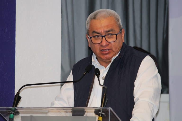 Hombre mayor con gafas y cabello gris, hablando en un micrófono durante una conferencia o evento formal.