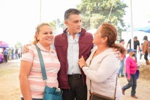 Persona conversando y sonriendo en un evento al aire libre, con fondo de gente y carpas en un día soleado.