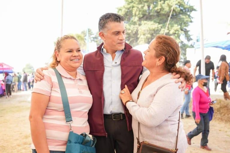 Persona conversando y sonriendo en un evento al aire libre, con fondo de gente y carpas en un día soleado.