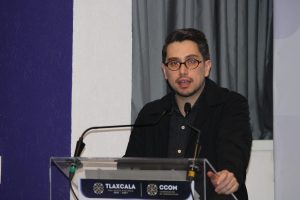 Persona hablando en un podio durante una conferencia en Tlaxcala, con fondo de cortinas grises y una pared blanca, vestido con chaqueta negra y gafas.