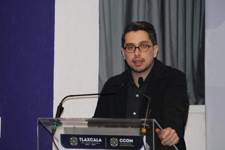 Persona hablando en un podio durante una conferencia en Tlaxcala, con fondo de cortinas grises y una pared blanca, vestido con chaqueta negra y gafas.