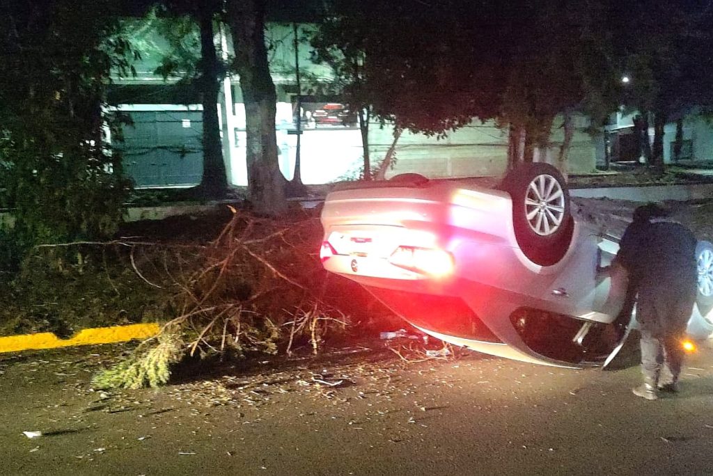 Un coche volcado sobre la calle, con daños visibles y luces traseras encendidas, cerca de árboles y un poste de luz en un área urbana durante la noche.
