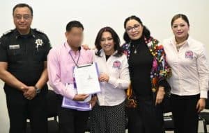 Grupo de personas en una ceremonia de reconocimiento, incluyendo un oficial de policía, un joven con certificado y tres mujeres, en un entorno de oficina o sala de reuniones.