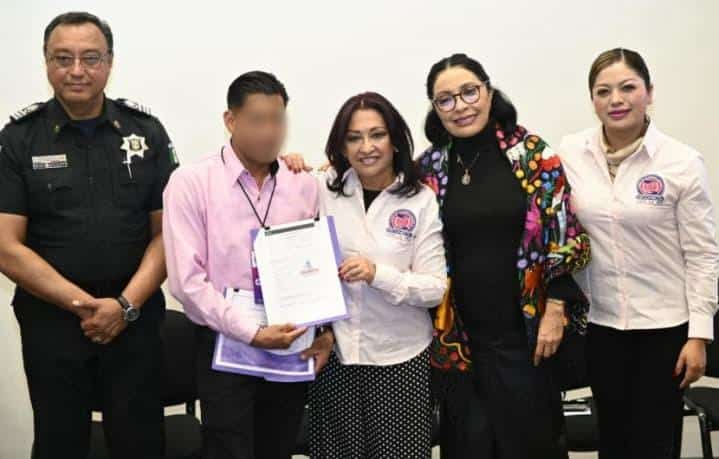 Grupo de personas en una ceremonia de reconocimiento, incluyendo un oficial de policía, un joven con certificado y tres mujeres, en un entorno de oficina o sala de reuniones.