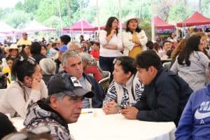 Personas participando en un evento al aire libre con muchas personas, algunos sentados en mesas y otros de pie, en un ambiente festivo con carpas y árboles de fondo.