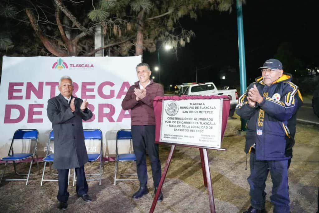 Personas en ceremonia de inauguración de obra pública en Tlaxcala, con un cartel que indica la entrega de obra en San Diego Metepec durante la noche.