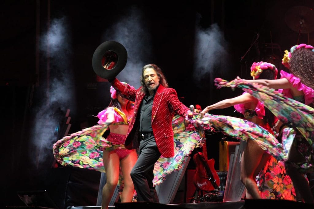 Un cantante con barba y cabello largo, vestido con saco rojo brillante, realiza una presentación en un escenario con bailarinas vestidas con trajes coloridos y flores, en un espectáculo en vivo.