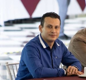 Persona joven con cabello oscuro y peinado corto, vistiendo una camisa azul, en un entorno moderno y profesional.