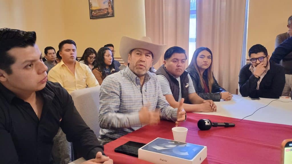 Asunto de reunión con varios jóvenes y un adulto en una mesa, en un entorno interior, en una sala con cortinas y una ventana al fondo, en un contexto de discusión o presentación.