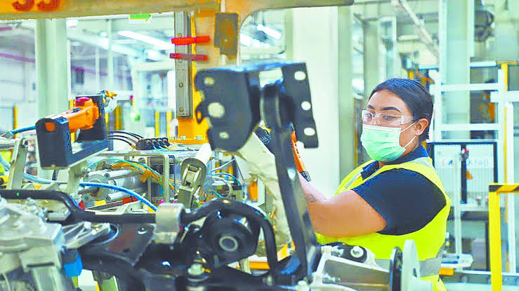 Trabajadora en línea de ensamblaje en una fábrica, usando equipo de protección y revisando componentes electrónicos.