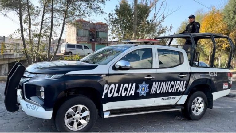 Policía municipal de Zacatelco en una patrulla negra y blanca, con un oficial en la parte trasera, en una calle con árboles y edificios al fondo, durante el día.
